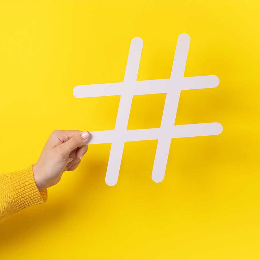 imagem de uma pessoa segurando o símbolo da hashtag branco com um parede de fundo na cor amarela, representando o assunto do artigo, que a agência de marketing digital criou.